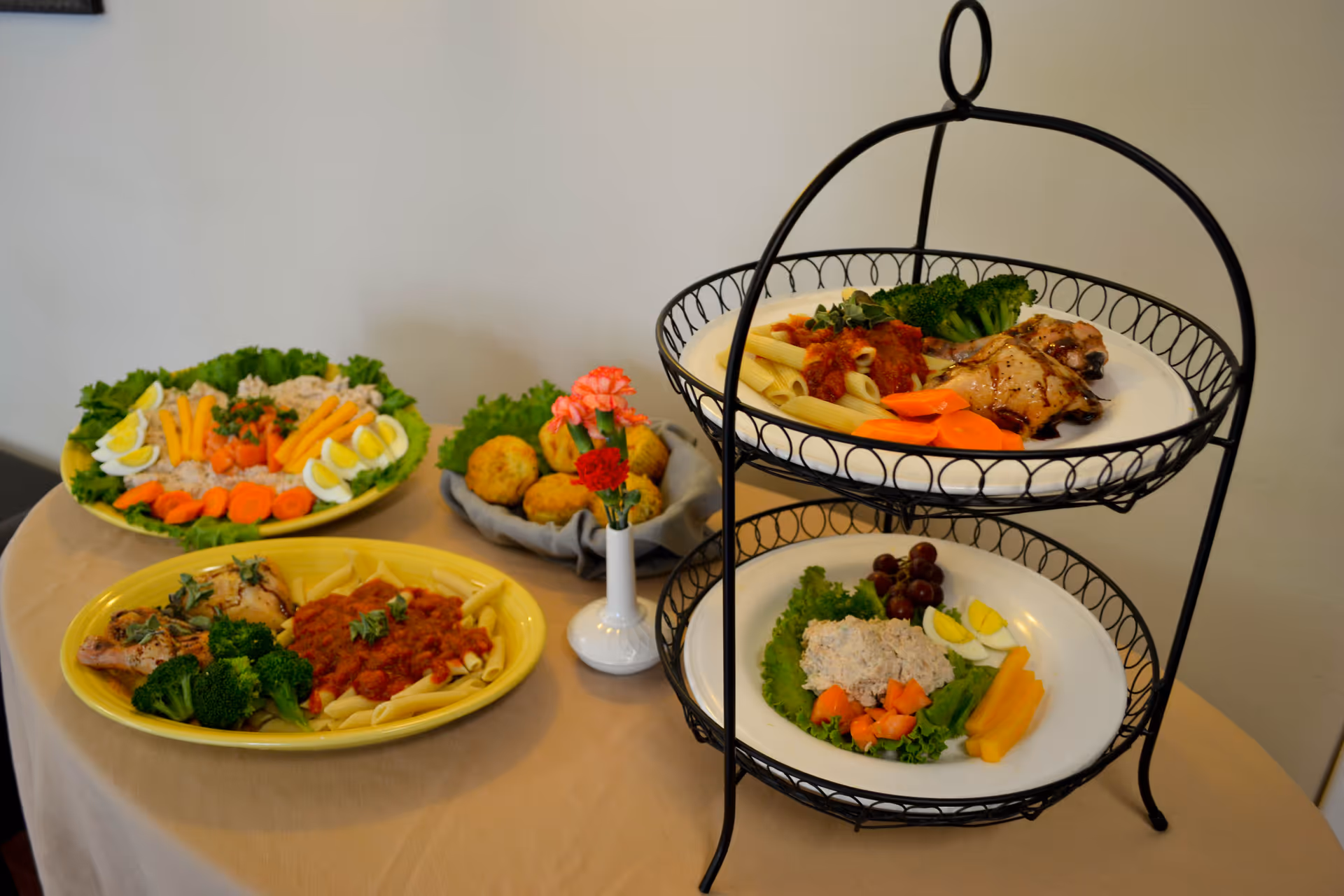 A table set with plates of food including pasta with tomato sauce, grilled chicken, broccoli, carrot slices, hard-boiled egg wedges, lettuce, tuna salad, grapes, and a basket of muffins, with a small vase holding pink and red flowers.