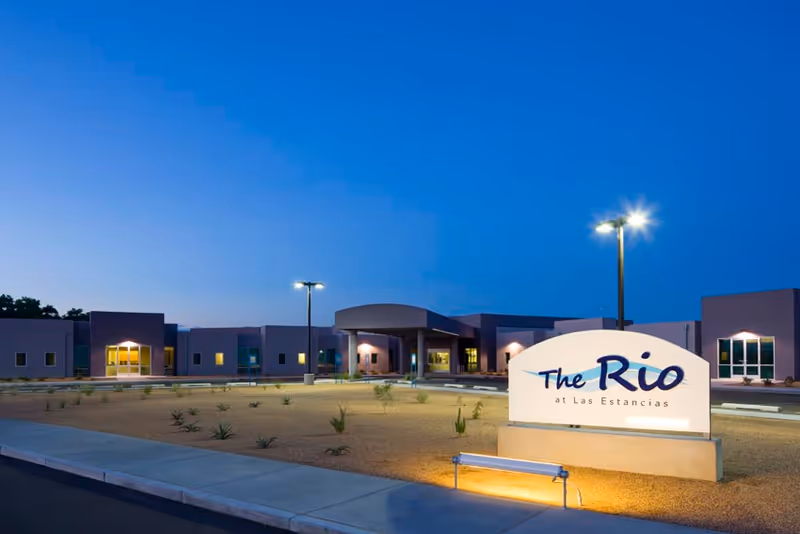 Dusk view of the front exterior of a senior living facility with a lit sign reading "The Rio at Las Estancias" in the foreground.
