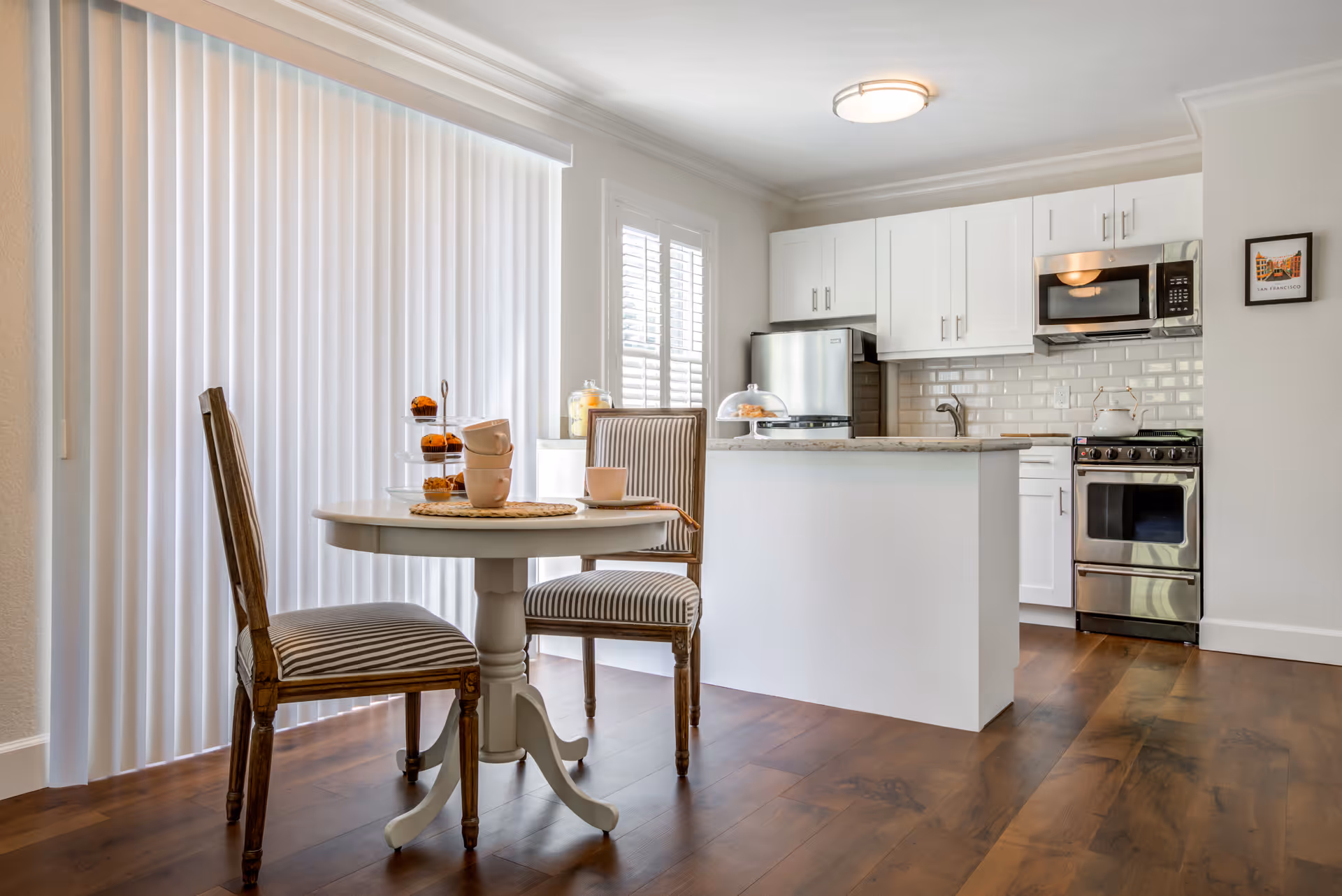 A bright kitchen and dining area featuring a small round table with two striped upholstered chairs. The kitchen has white cabinets, a stainless steel refrigerator, stove, and microwave. The floor is wooden, and there are vertical blinds covering a large window or sliding door.