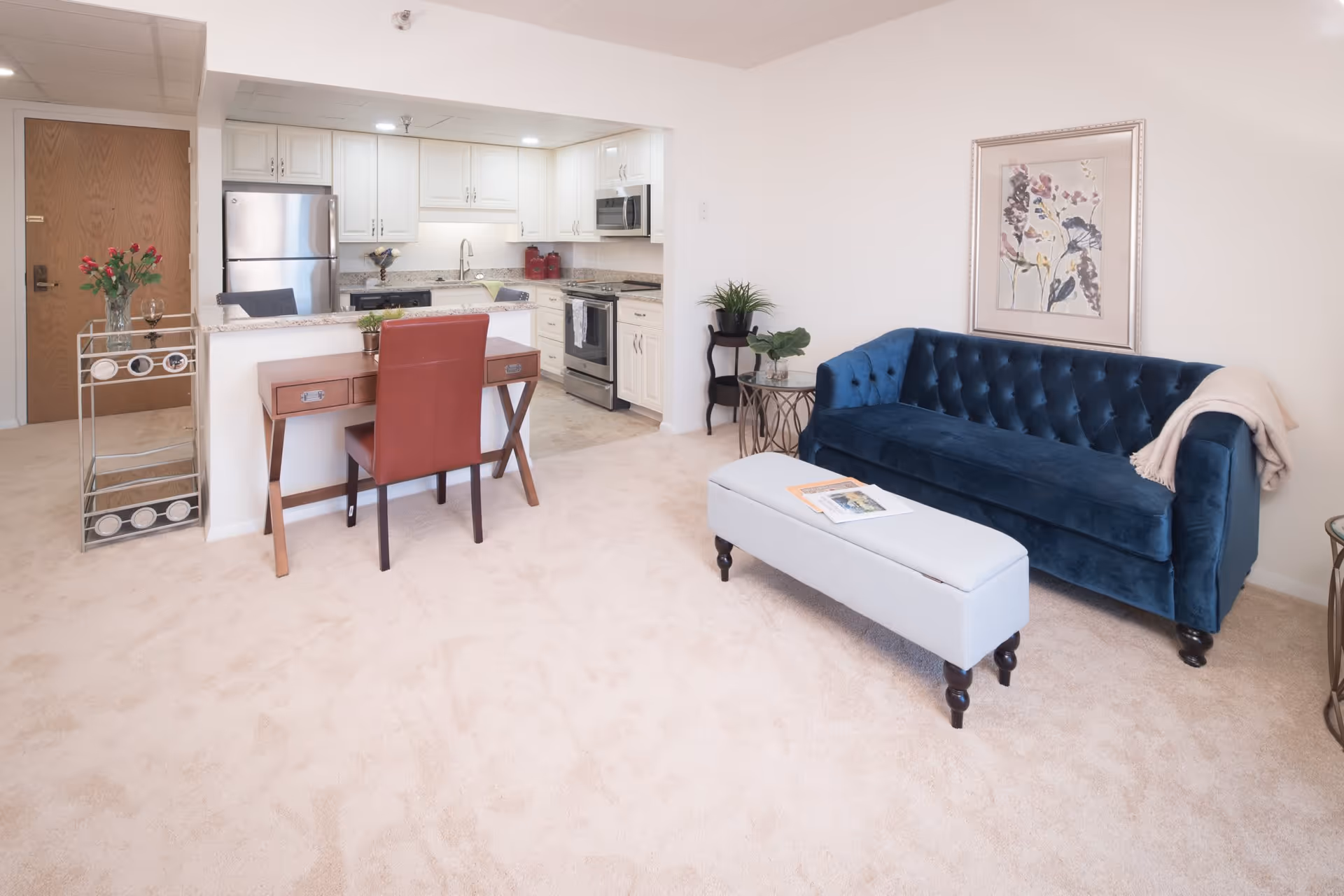 A bright and clean living area with beige carpet, featuring a dark blue tufted sofa with a light beige throw blanket, a light gray upholstered bench with magazines on top, a small side table with a plant, and a wooden desk with a red chair. In the background, there is an open kitchen with white cabinets, stainless steel appliances, and a granite countertop. A wooden door and a small metal cart with a vase of red flowers and a wine glass are also visible.