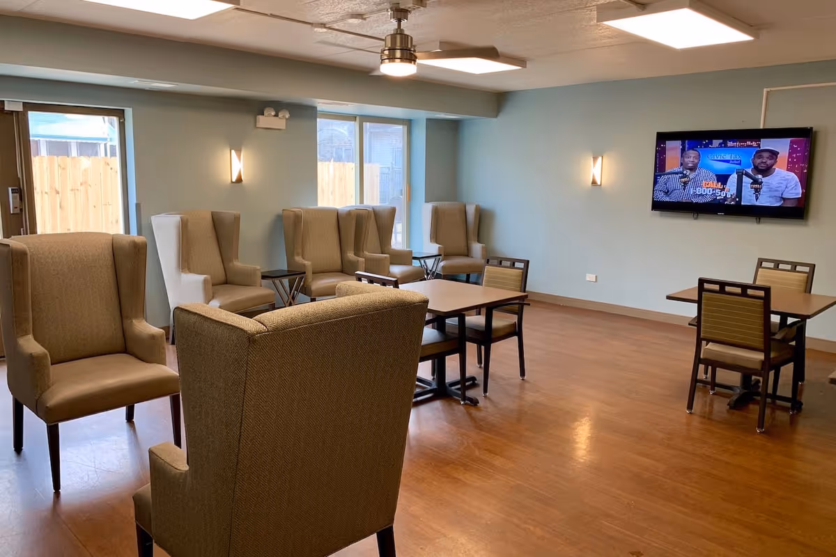 A common area in a senior living facility with several beige armchairs and tables with chairs on a wooden floor. The walls are painted light blue, and there are two windows and a glass door letting in natural light. A flat-screen TV is mounted on the wall showing two men speaking.