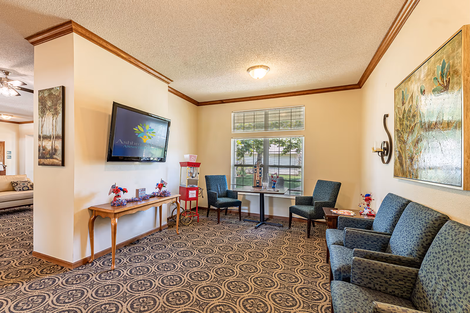A cozy living room area in Ashbrook Village assisted living facility featuring patterned carpet, a wall-mounted TV displaying the Ashbrook Village logo, a wooden console table decorated with patriotic items, a red popcorn machine, two blue upholstered chairs around a small table near a window, and a blue upholstered sofa with a large framed painting on the wall.