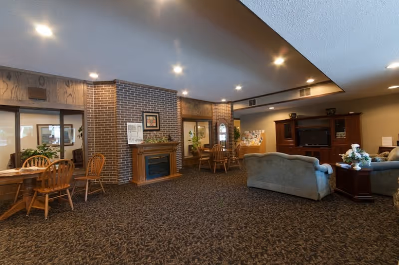 A spacious senior living community common area featuring a carpeted floor, a brick fireplace with a wooden mantel, several wooden tables and chairs, a large wooden entertainment center with a television, and comfortable upholstered sofas. The room is well-lit with recessed ceiling lights and decorated with plants and framed artwork.