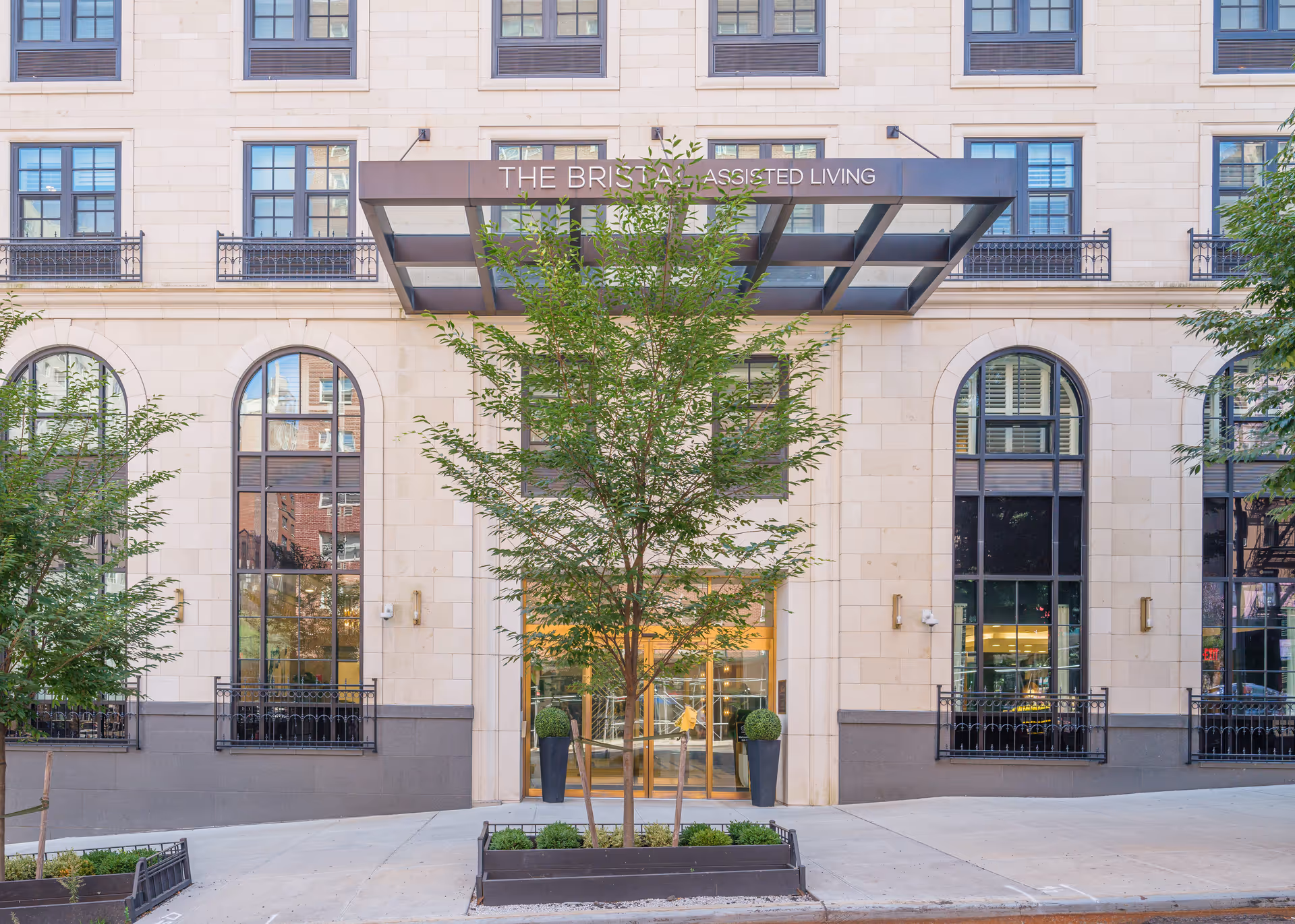 Front exterior view of The Bristal Assisted Living at York Avenue building with large arched windows, a glass and metal canopy above the entrance, and small trees planted in front.