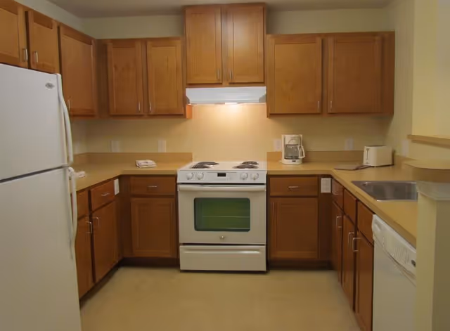 A kitchen with wooden cabinets, a white refrigerator on the left, a white electric stove with oven in the center, a coffee maker and toaster on the right countertop, a stainless steel sink, and a dishwasher underneath the counter on the right side.