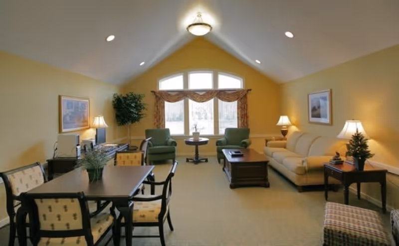 A cozy living room with yellow walls and a vaulted ceiling featuring recessed lighting and a central ceiling light. The room includes a beige sofa, two green armchairs, a wooden coffee table, and a dining table with six chairs. There are two table lamps on side tables, framed artwork on the walls, a potted plant near the window, and a large window with patterned curtains letting in natural light.