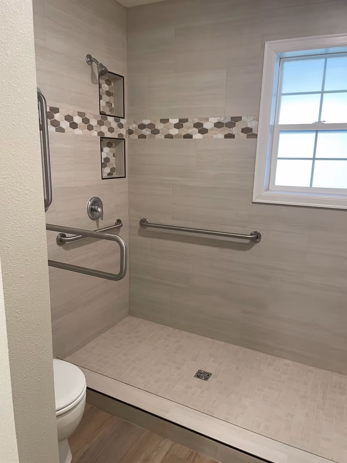 A modern accessible shower area with beige tiles and a horizontal strip of hexagonal patterned tiles. The shower has two built-in niches for toiletries, stainless steel grab bars on the walls, and a handheld showerhead. There is a window with frosted glass on the right side and a toilet partially visible on the left.