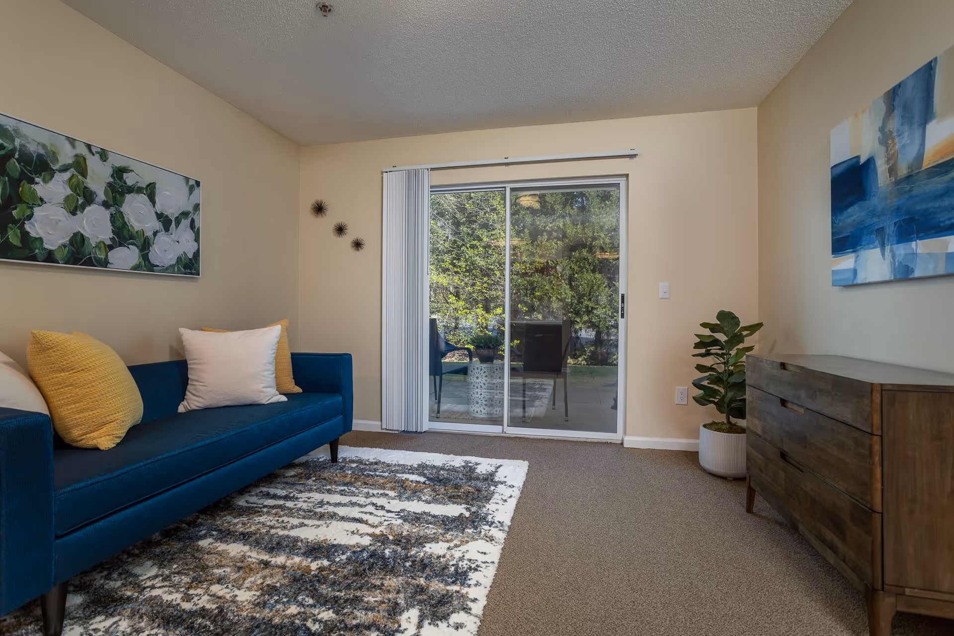 A cozy living room with a blue sofa adorned with yellow and white pillows, a patterned rug on the carpeted floor, a wooden dresser with a potted plant beside it, abstract and floral paintings on the walls, and a sliding glass door leading to an outdoor patio with chairs and greenery visible outside.
