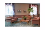A cozy living room area in a senior living facility with three upholstered armchairs and a matching sofa arranged around a wooden coffee table with a plant centerpiece. The room has large windows with patterned curtains, framed pictures on the wall, and a tall potted plant near the window.