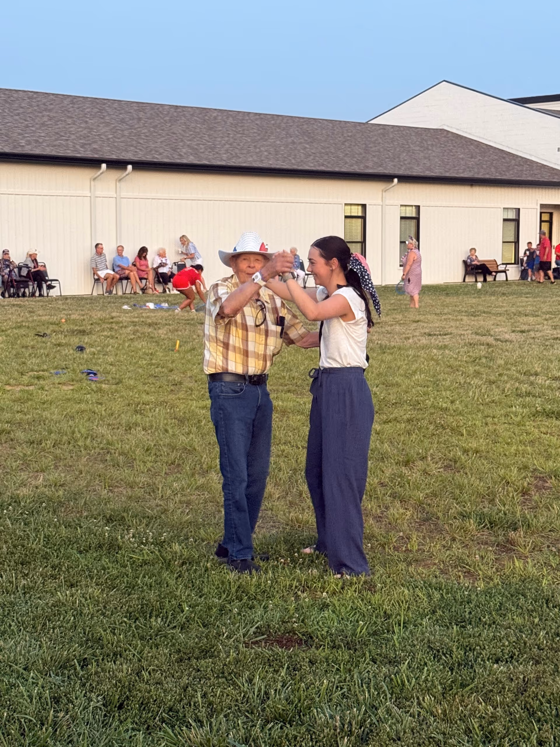 An elderly man wearing a white cowboy hat and a plaid shirt dances with a young woman dressed in a white top and blue pants on a grassy lawn outside a building. In the background, several people are sitting on chairs and benches near the building, enjoying the outdoor setting.