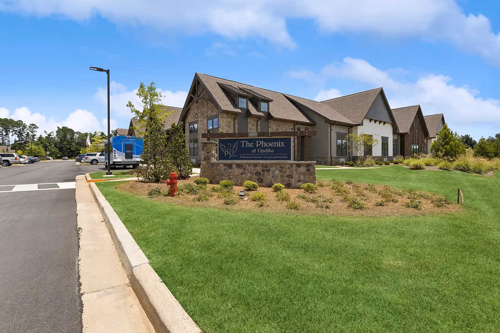 Exterior view of The Phoenix at Opelika senior living community building with a stone and wood facade, a landscaped lawn, and a parking lot with several vehicles under a blue sky with some clouds.