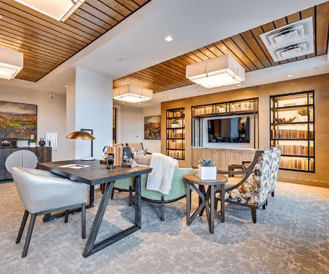 Spacious lounge with seating, tables, built-in shelving and a wall-mounted TV under a wood-paneled ceiling.