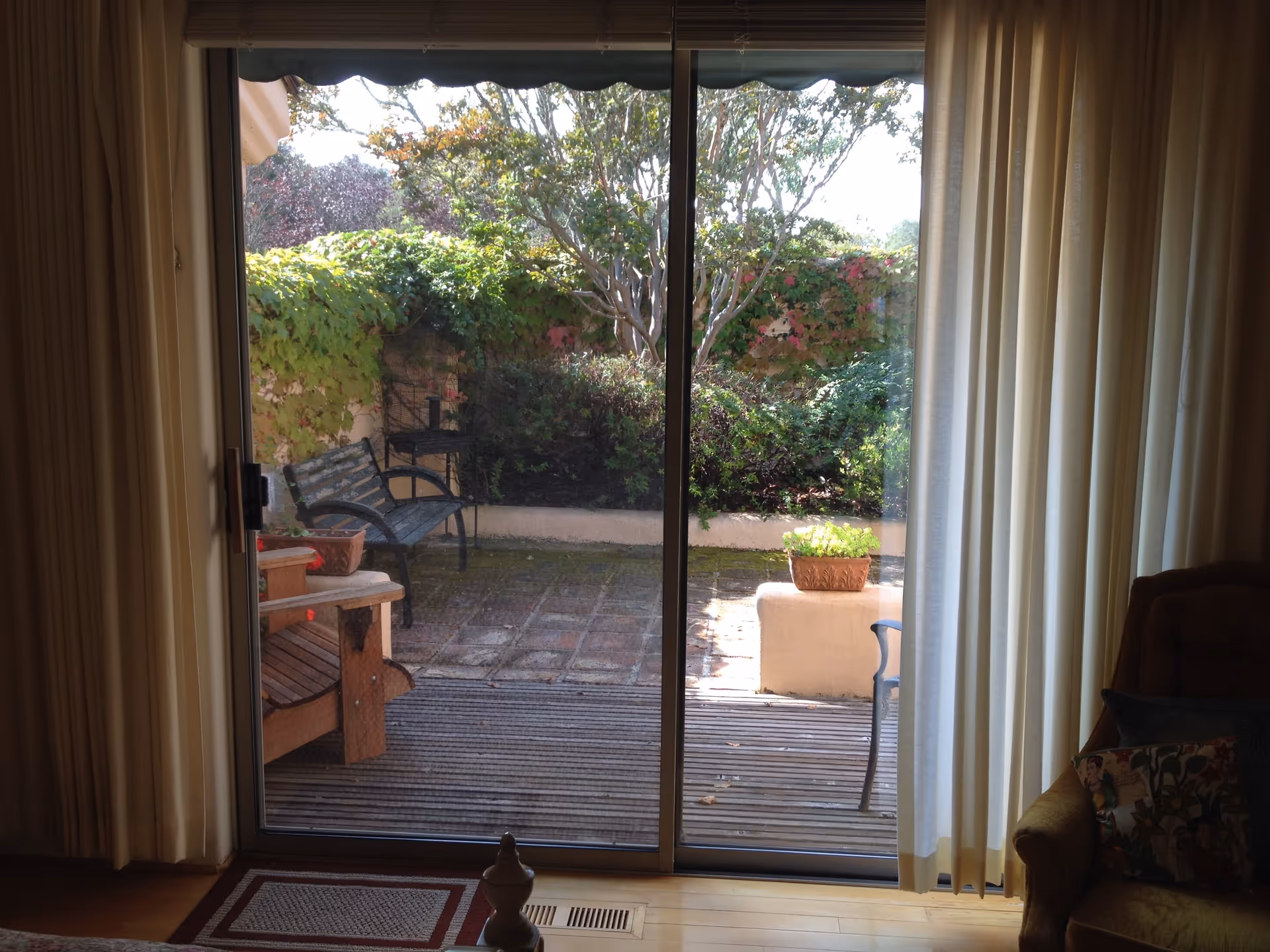 View through a sliding glass door from an interior room to an outdoor patio area with wooden benches, a small table, potted plants, and lush greenery including bushes and a tree. Light curtains frame the door and part of an armchair with patterned pillows is visible inside.