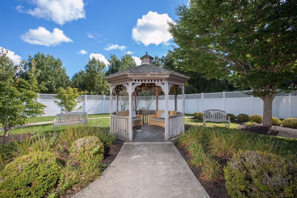 Outdoor garden area with a wooden gazebo featuring cushioned seating inside. The gazebo is surrounded by green grass, bushes, trees, and two wooden benches. A white fence encloses the area under a blue sky with scattered clouds.