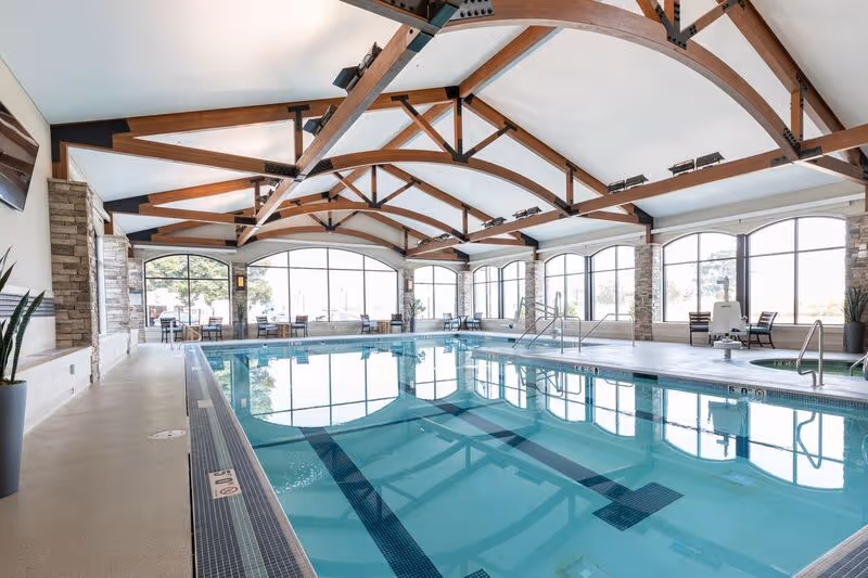 Indoor swimming pool area with clear blue water, surrounded by large windows letting in natural light. The ceiling features wooden beams with metal supports, and there are chairs and tables along the sides of the pool. The space has a clean, modern design with stone pillars and potted plants.