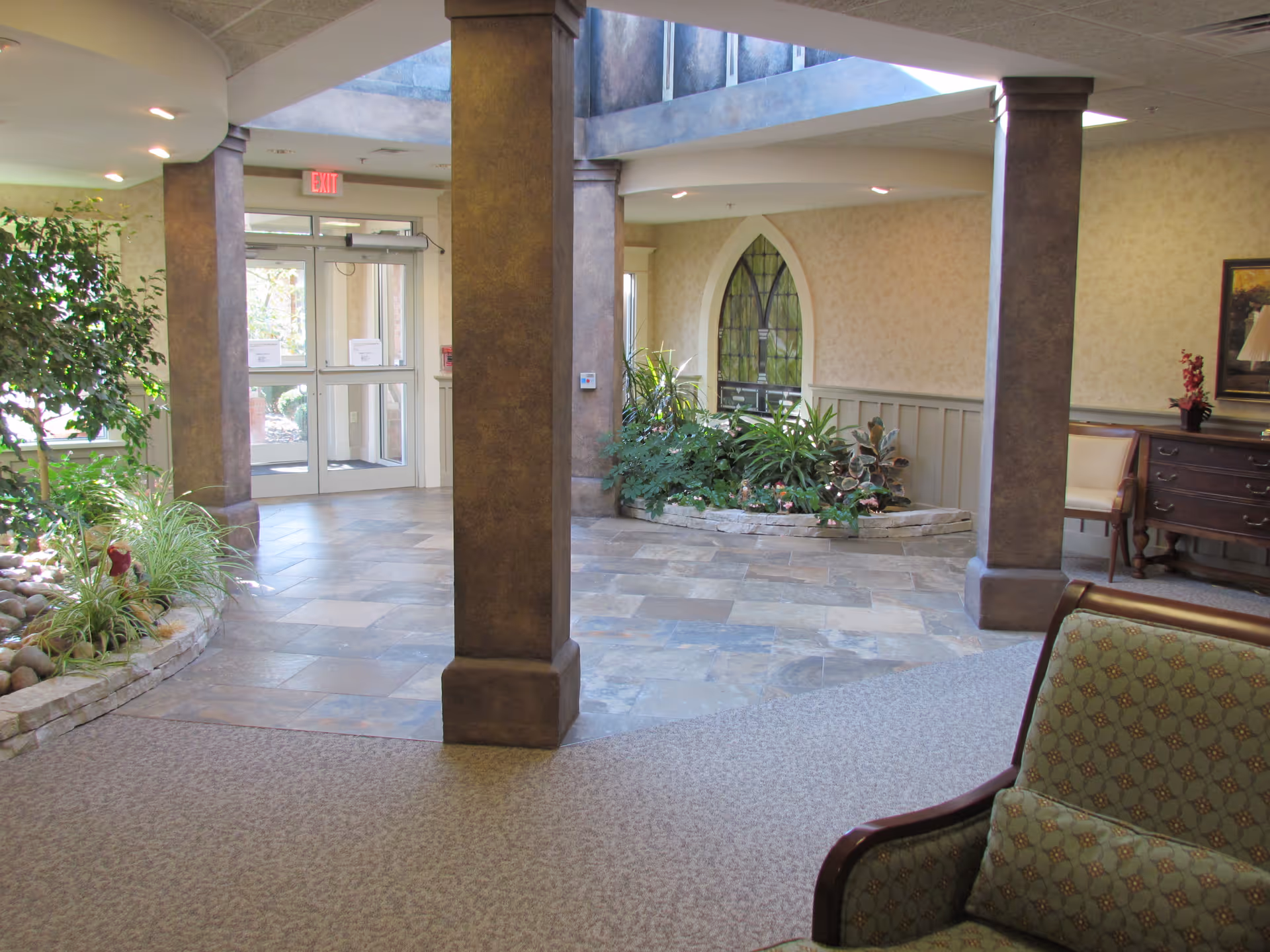 Interior view of a senior living facility lobby area with stone tile flooring, carpeted seating area with green patterned armchairs, decorative indoor plants in stone-bordered planters, large pillars, and a glass exit door leading outside.