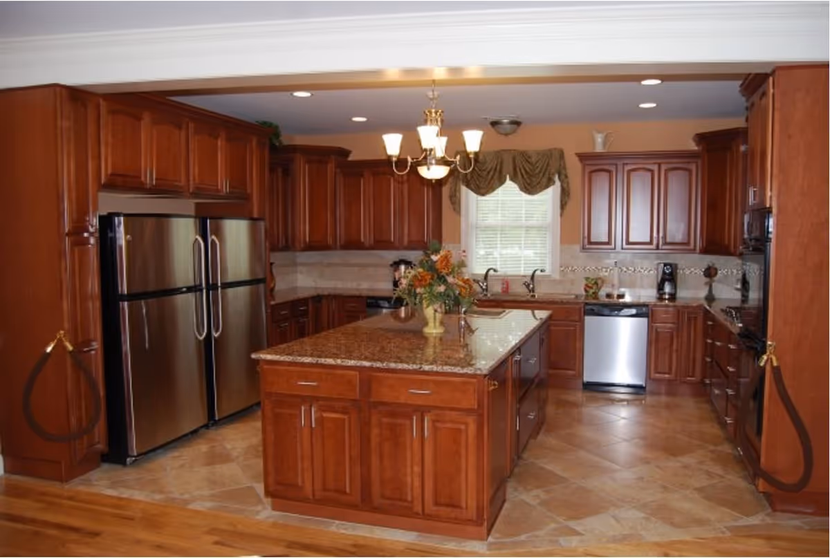 Spacious kitchen with wooden cabinets, a large central island with a granite countertop, stainless steel refrigerator, dishwasher, and oven. A window with a valance is above the sink, and a chandelier hangs from the ceiling.