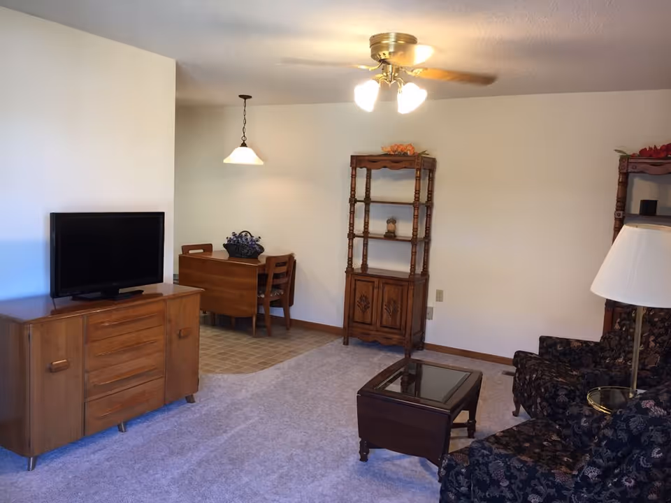 A living room area with a wooden TV stand holding a flat-screen television on the left, a small dining table with two chairs in the background under a hanging light, a wooden shelving unit against the wall, a glass-top coffee table, two patterned armchairs, and a floor lamp with a white lampshade. The room has light-colored walls, carpeted flooring, and a ceiling fan with lights.