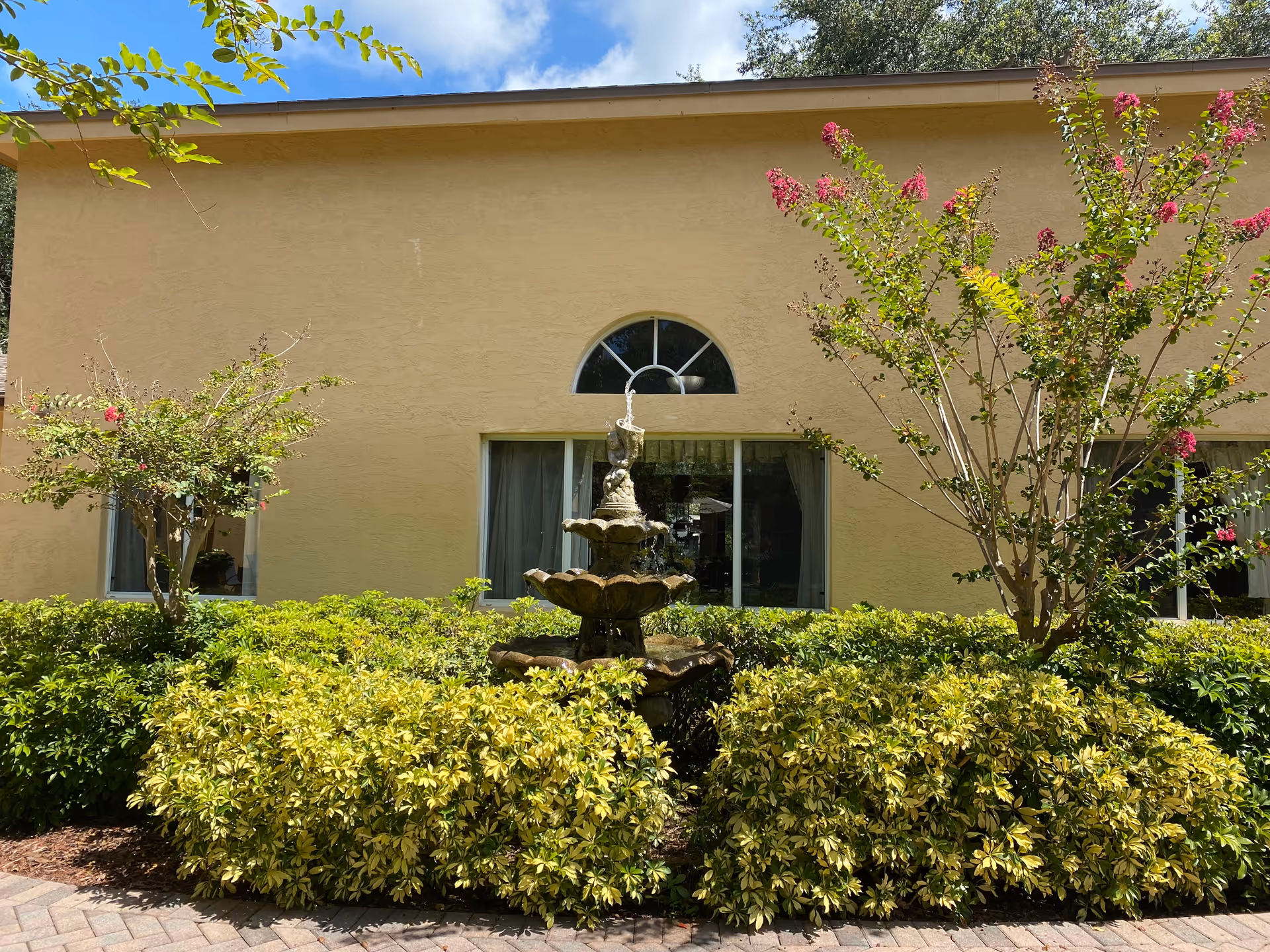 Small courtyard fountain surrounded by shrubs and flowering trees in front of a beige building with arched and rectangular windows.