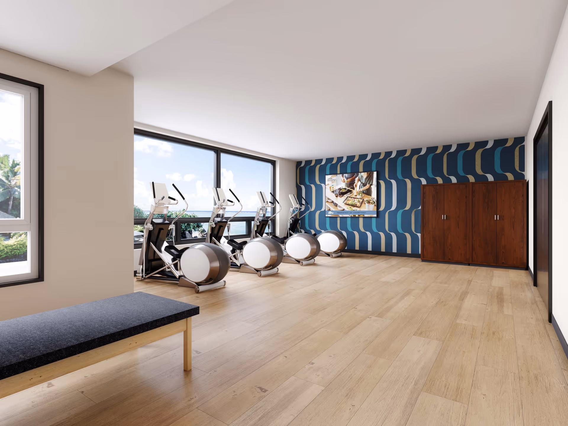 Bright fitness room with exercise bikes by large windows, a patterned blue accent wall, wooden storage cabinets and a bench on a light wood floor.