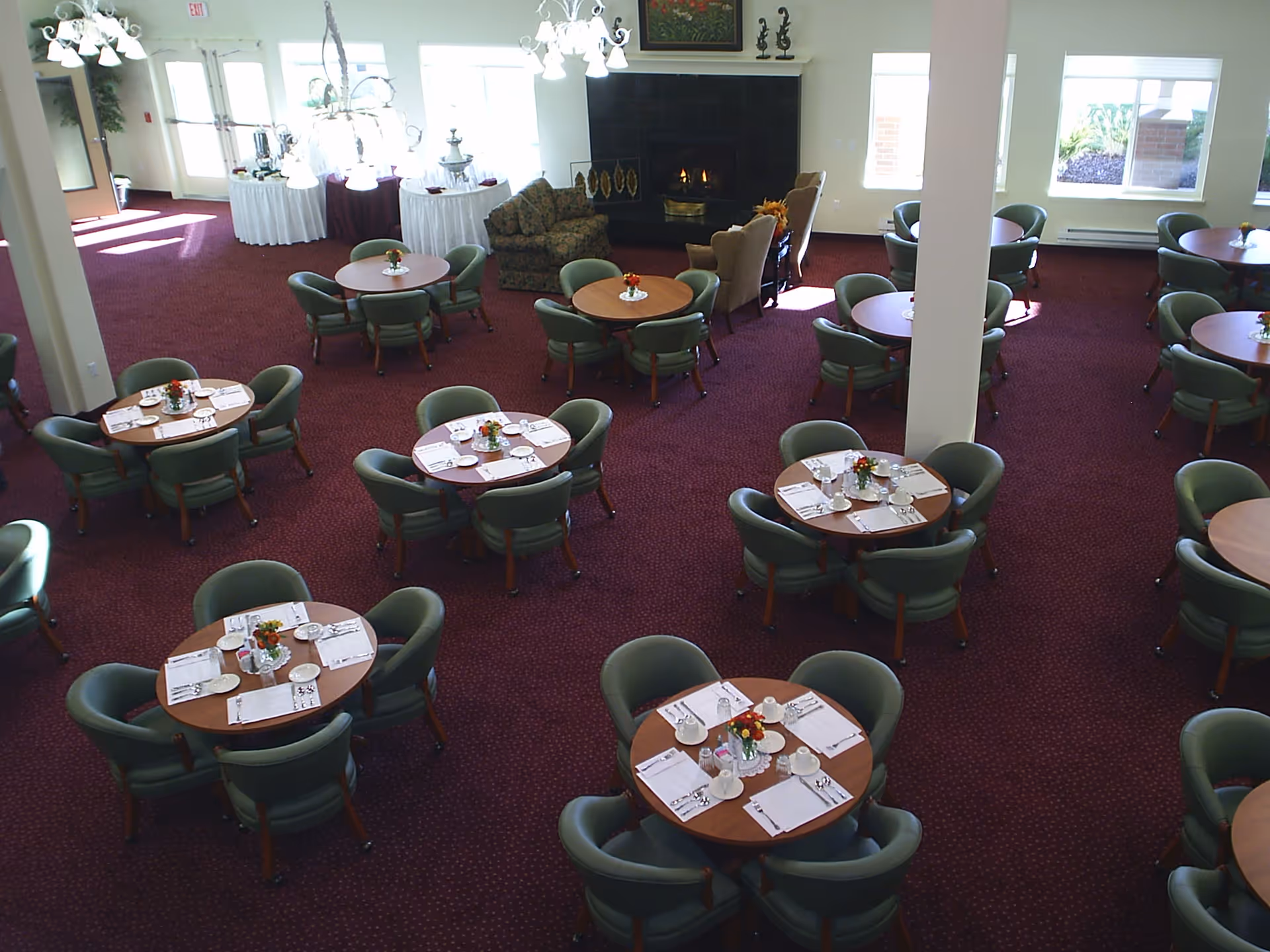 A spacious dining room with multiple round tables each set with placemats, cups, and utensils. The room has green cushioned chairs around the tables, a red carpeted floor, large windows letting in natural light, and a fireplace with seating near it. There are chandeliers hanging from the ceiling and a buffet table in the background.