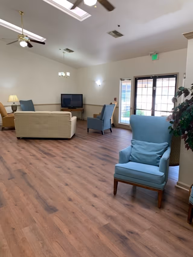 Bright common living room with blue armchairs, sofas, a television, and wood-look flooring.
