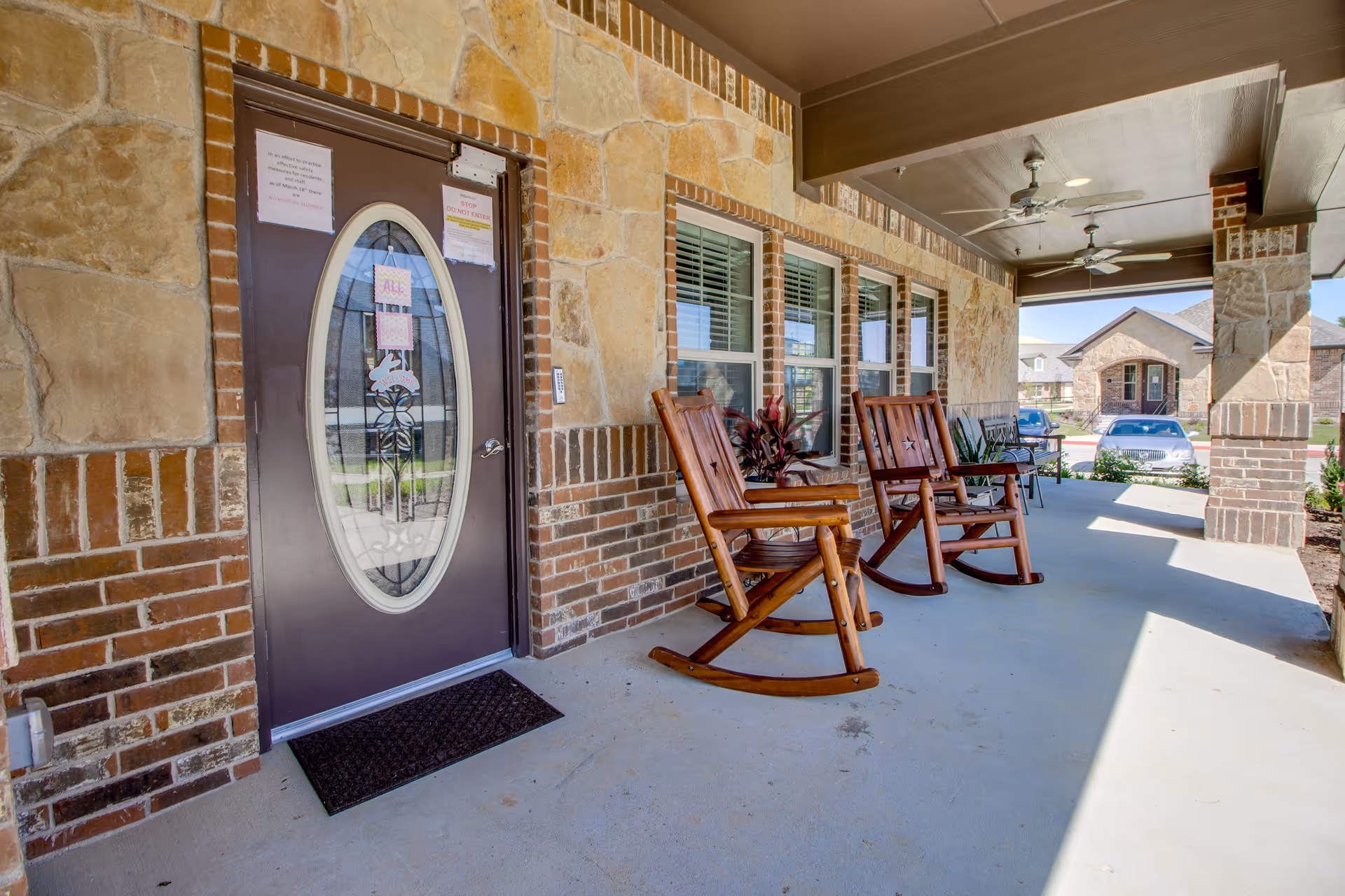 Covered porch area of a senior living facility with two wooden rocking chairs, a bench, potted plants, and a brown door with an oval glass window. The porch has stone and brick walls, ceiling fans, and overlooks a parking area with cars and other buildings in the background.