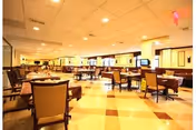 A spacious dining room with multiple tables and chairs arranged neatly. The room has a checkered tile floor, warm lighting from ceiling fixtures, and windows along the far wall. The tables are set with placemats and some have small centerpieces.