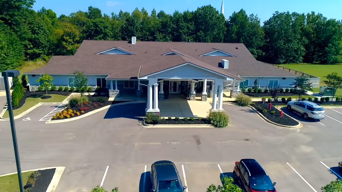 Front exterior view of BeeHive Homes of Collierville, a single-story building with a large covered entrance supported by white columns, surrounded by a parking lot with several cars and landscaped greenery, with trees in the background.