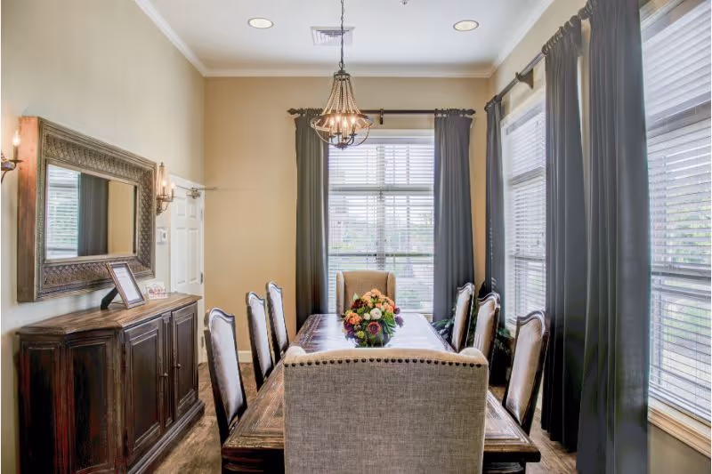 A well-lit dining room with a long wooden table surrounded by eight upholstered chairs. A floral centerpiece is placed on the table. The room has large windows with dark curtains, a chandelier hanging from the ceiling, a large decorative mirror on the wall, and a wooden sideboard beneath the mirror.