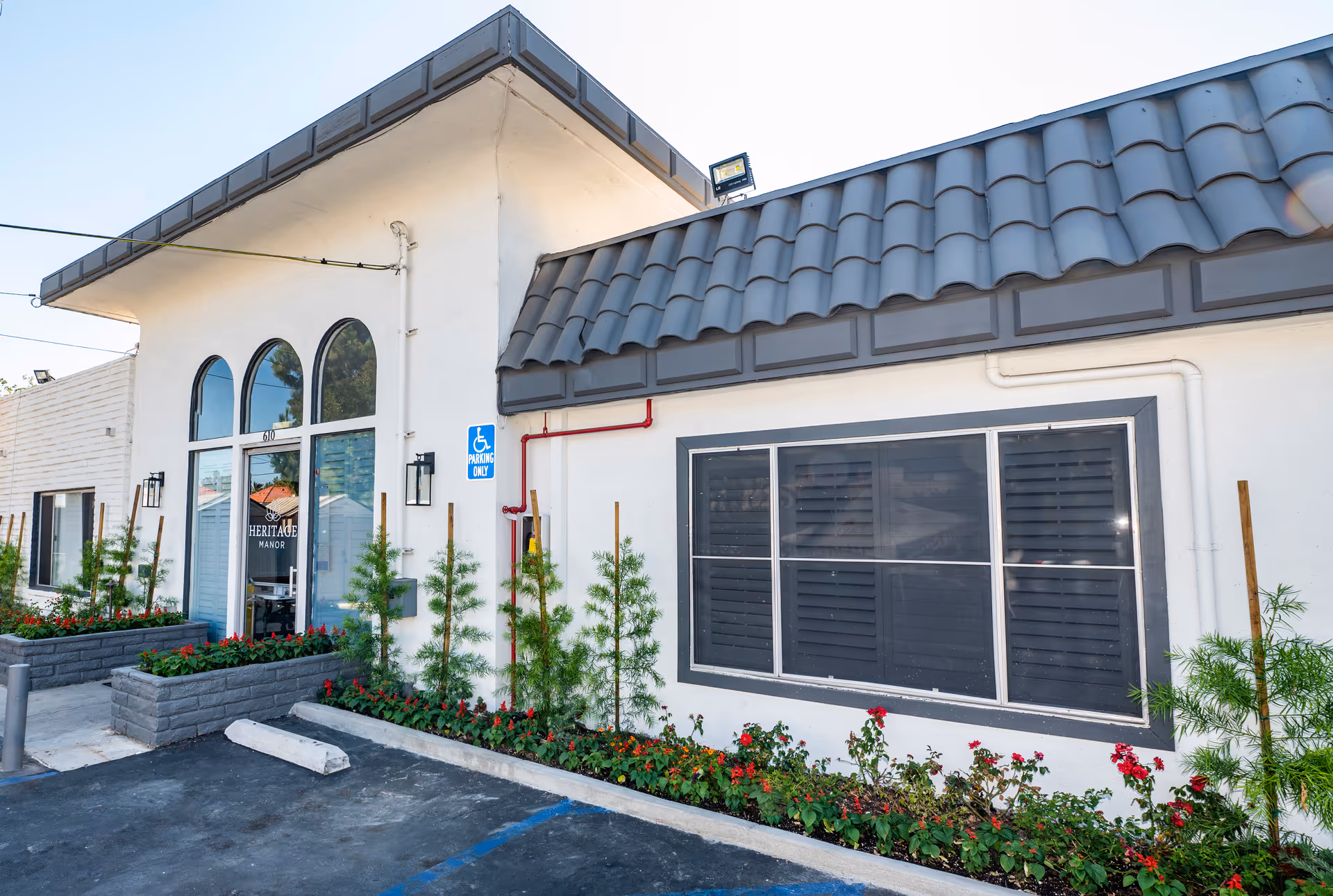 Exterior view of Heritage Manor 頤康療養院 showing the building entrance with large arched windows and a door with the facility's logo. There are flower beds with green plants and red flowers along the front, a handicapped parking sign, and a tiled roof section above the window.