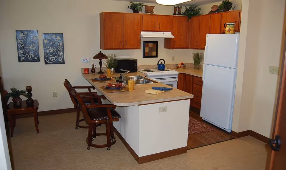 A compact kitchen area in a senior living facility featuring wooden cabinets, a white refrigerator, a white stove with a blue kettle on it, and a countertop with a sink. The countertop extends into a breakfast bar with two wooden chairs. The kitchen is decorated with plants, a lamp, and framed artwork on the wall.