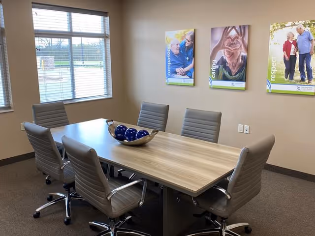 A conference room with a rectangular wooden table surrounded by six gray office chairs on wheels. On the table is a decorative bowl with blue spheres. The room has beige walls with three framed posters displaying words 'compassion', 'caring', and 'respect' along with images of elderly people. Two windows with blinds allow natural light into the room.