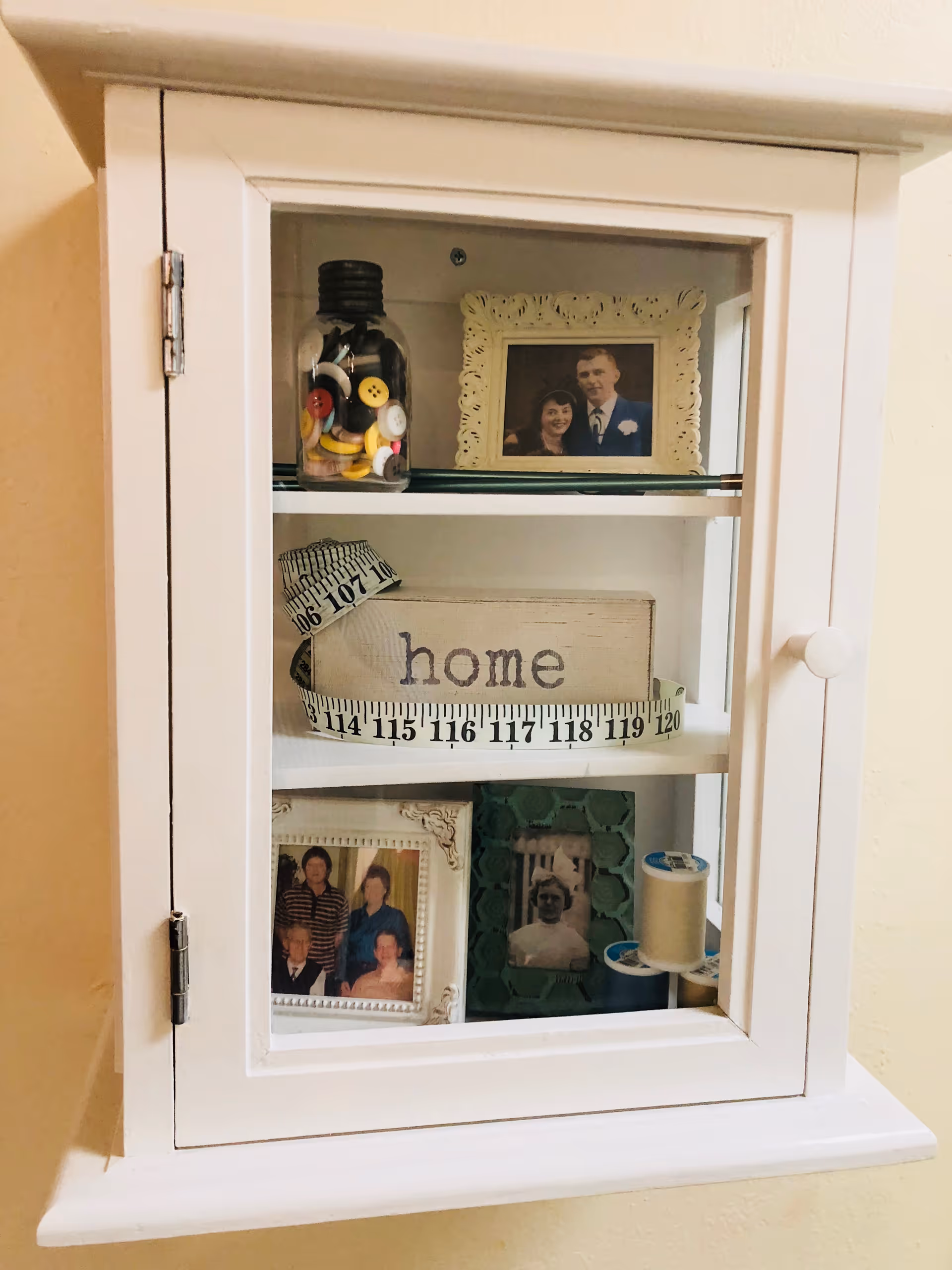 Wall-mounted white display cabinet with framed photos, a wooden 'home' sign, jar of buttons and sewing supplies behind a glass door.
