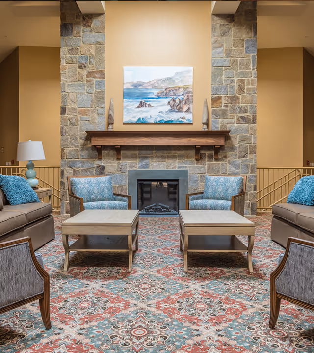 A cozy living room with a stone fireplace centered on the back wall, topped with a wooden mantel and a painting of a coastal scene. The room features two blue patterned armchairs and two beige sofas with blue throw pillows arranged around two wooden coffee tables on a colorful patterned rug. A lamp with a blue base is visible on the left side.