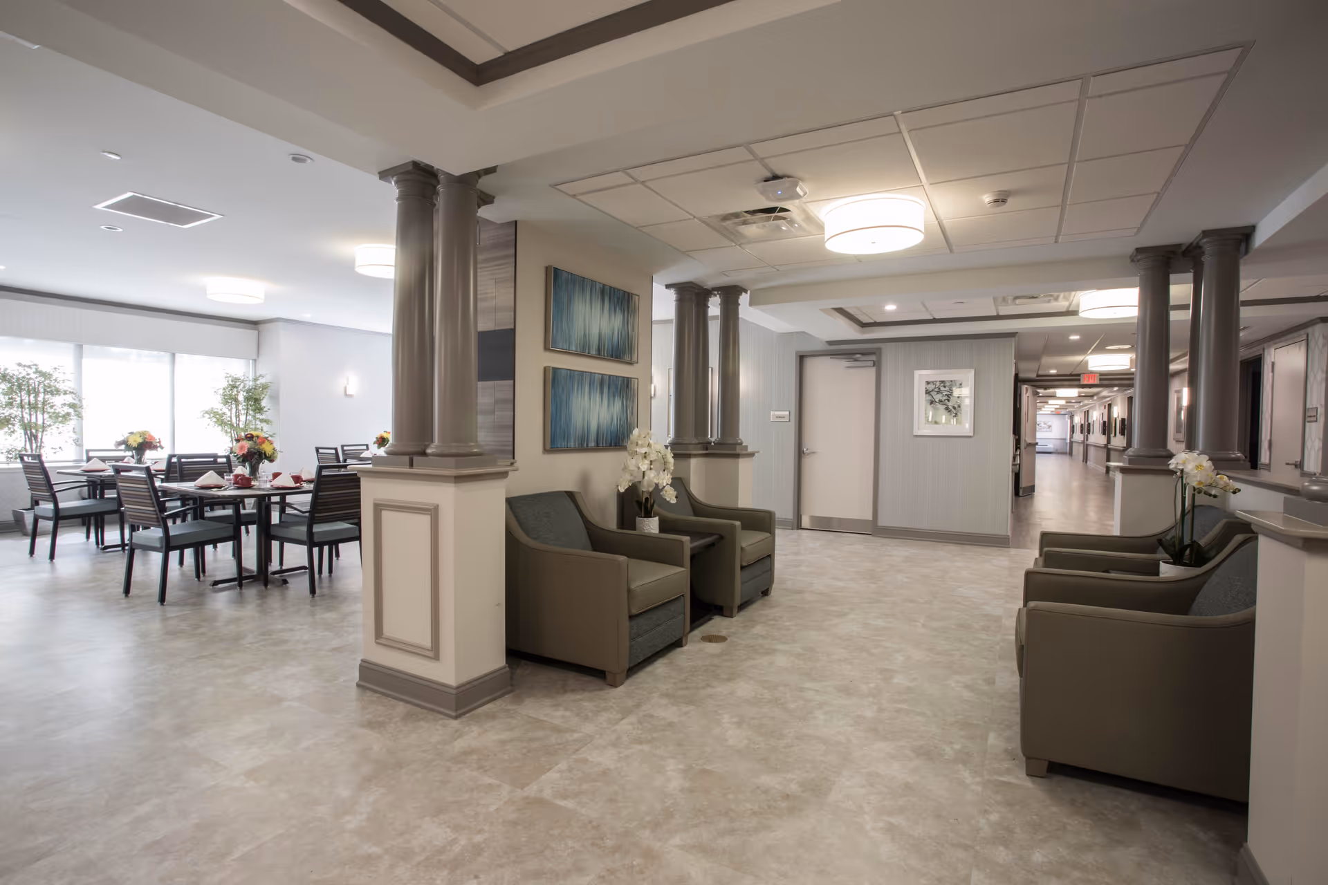 Interior view of a senior living facility showing a seating area with armchairs and decorative columns, adjacent to a dining area with tables and chairs set with flowers and napkins. The space is well-lit with ceiling lights and natural light from large windows.