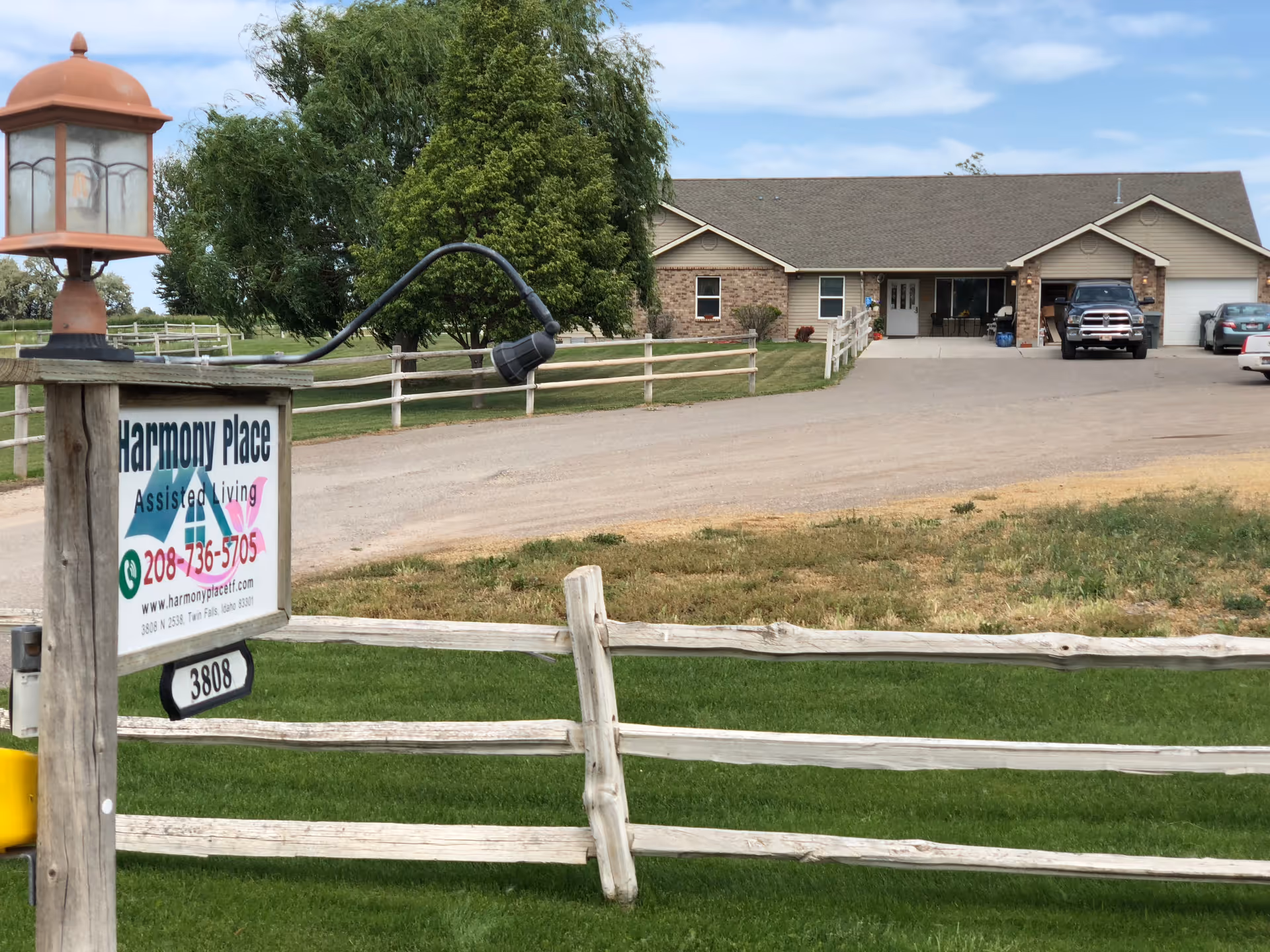 Exterior view of Harmony Place Assisted Living facility showing a single-story building with a driveway, parked vehicles, and a wooden fence with a sign displaying the facility's name, phone number, and website. There is a lamp post attached to the sign and green grass in the foreground.