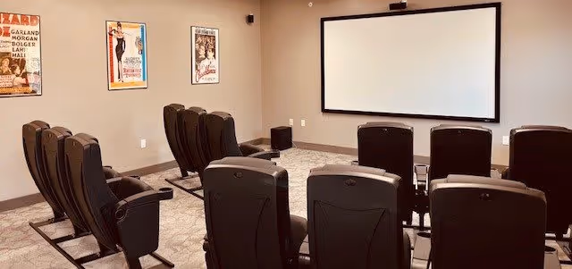 A small theater room with two rows of black reclining chairs facing a large white projection screen mounted on a beige wall. The room has carpeted flooring and three vintage movie posters hanging on the left wall.