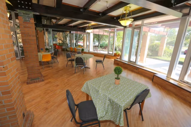 A spacious dining area with wooden floors and large windows letting in natural light. Several tables covered with green patterned tablecloths are arranged throughout the room, each with a small potted plant centerpiece. Black and orange chairs are placed around the tables. The room features exposed beams on the ceiling and brick pillars along one side.
