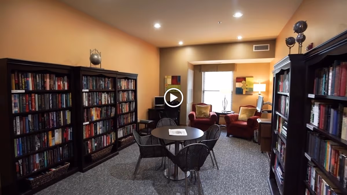 A cozy communal library room with tall bookshelves lining the walls, a round table with wicker chairs in the center, and two upholstered armchairs by a window.