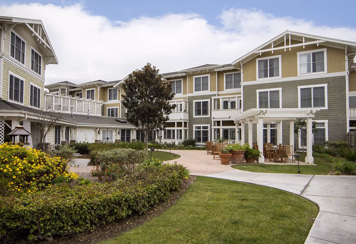Exterior view of a multi-story senior living building with a landscaped courtyard, pergola, and outdoor seating.