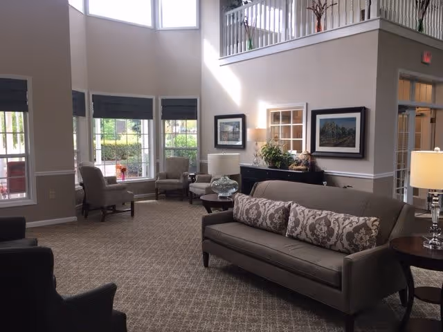 A bright and spacious living room area with large windows letting in natural light. The room features a gray sofa with patterned cushions, several armchairs, a side table with a lamp, framed artwork on the walls, and decorative plants. The space has a high ceiling and a carpeted floor with a subtle pattern.