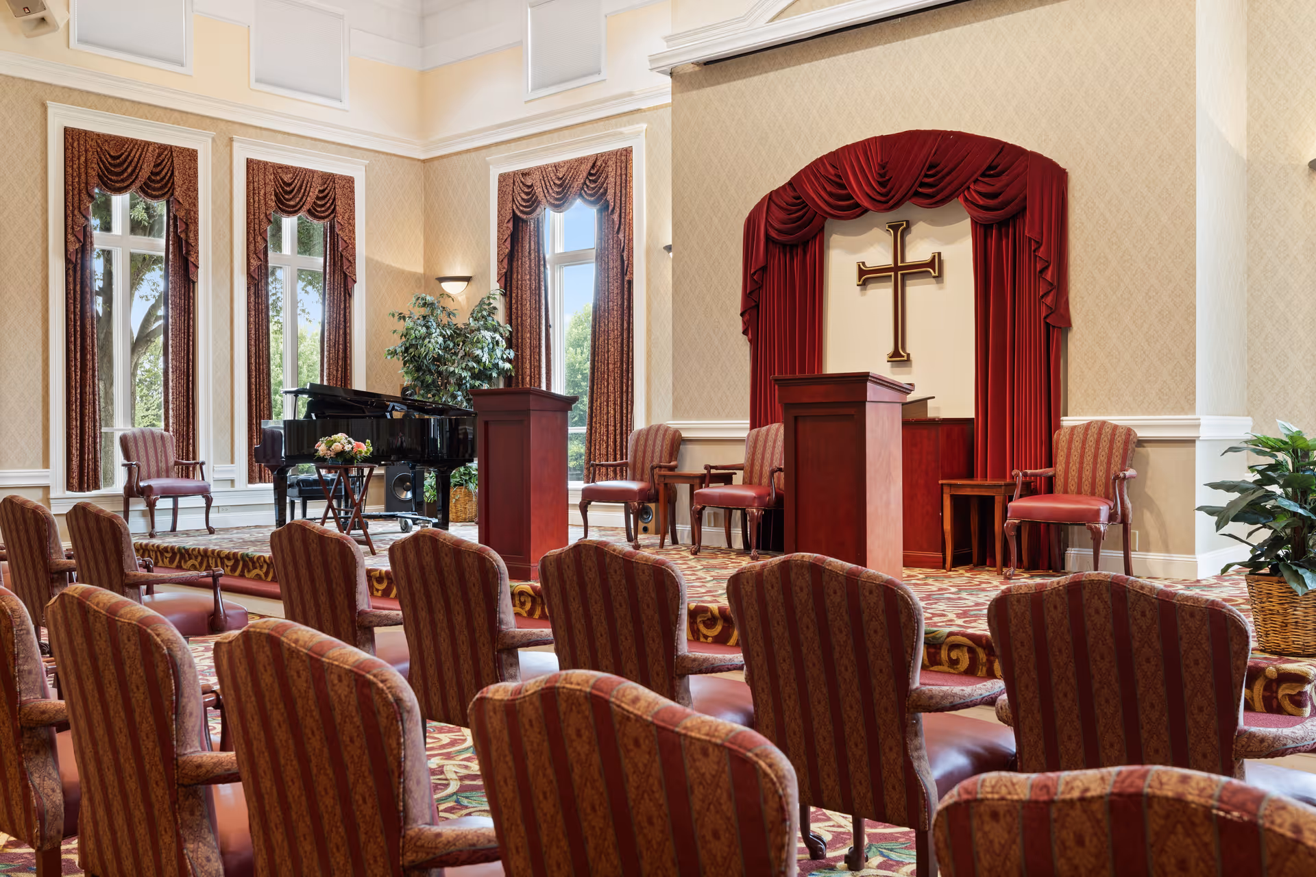 Interior chapel or assembly room with rows of upholstered chairs facing a podium, piano, and a stage with a cross framed by red curtains.
