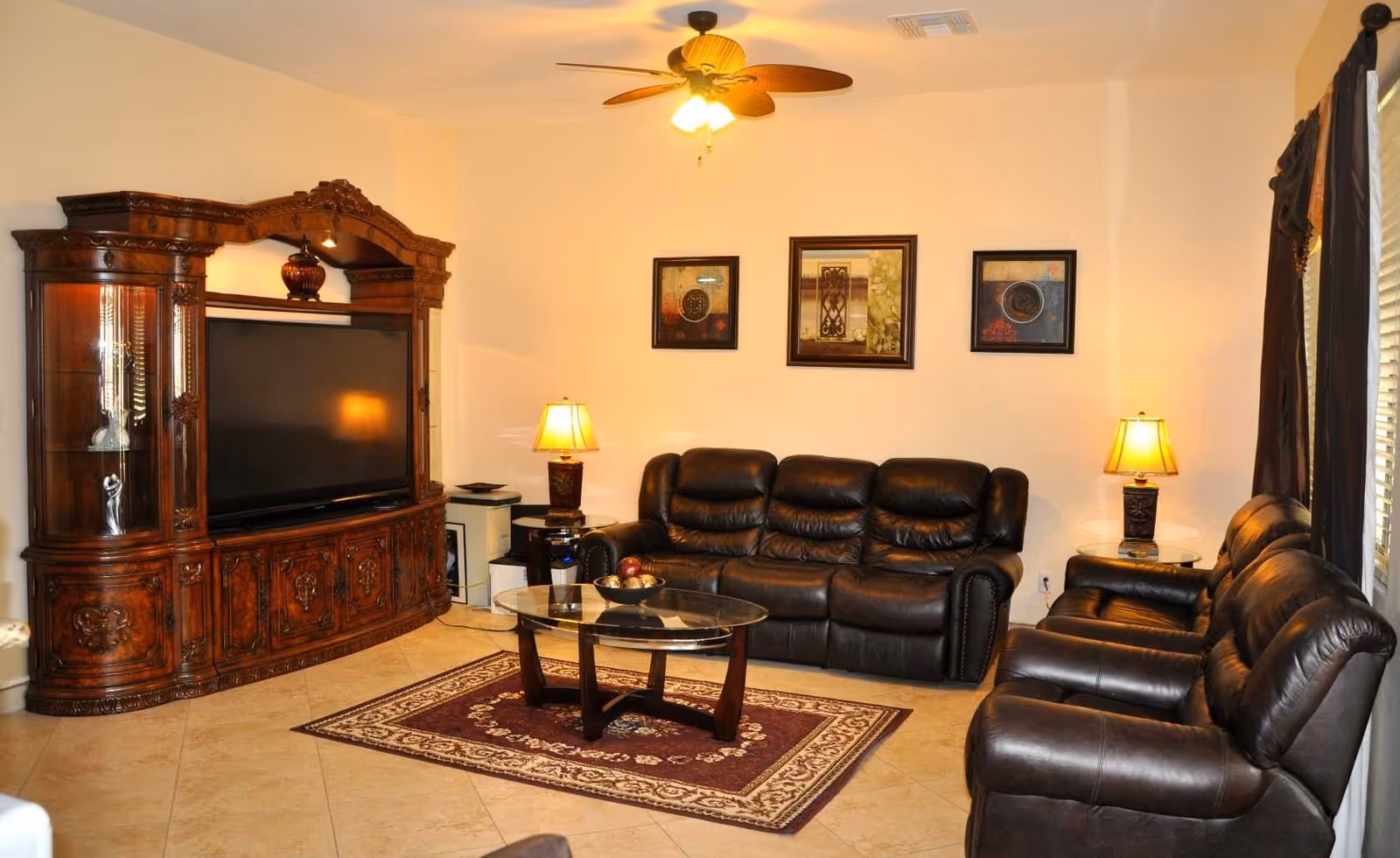 A living room with a large ornate wooden entertainment center holding a flat-screen TV, a dark brown leather sofa, two matching leather armchairs, a glass coffee table on a patterned rug, two table lamps on side tables, three framed pictures on the wall, and a ceiling fan with a light fixture.
