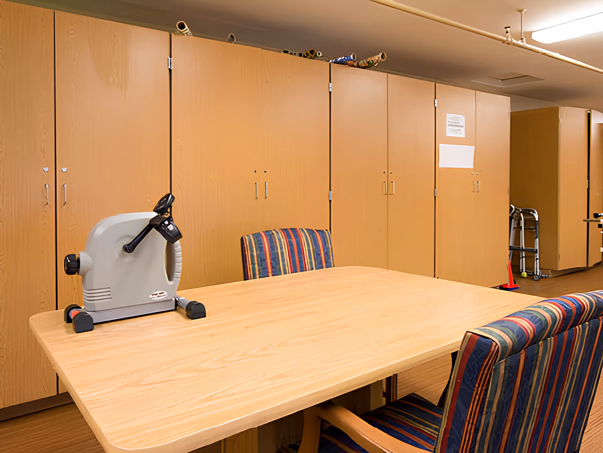 Interior activity room with a wooden table and striped chairs, a small tabletop exercise machine on the table, and tall wooden storage cabinets along the wall.