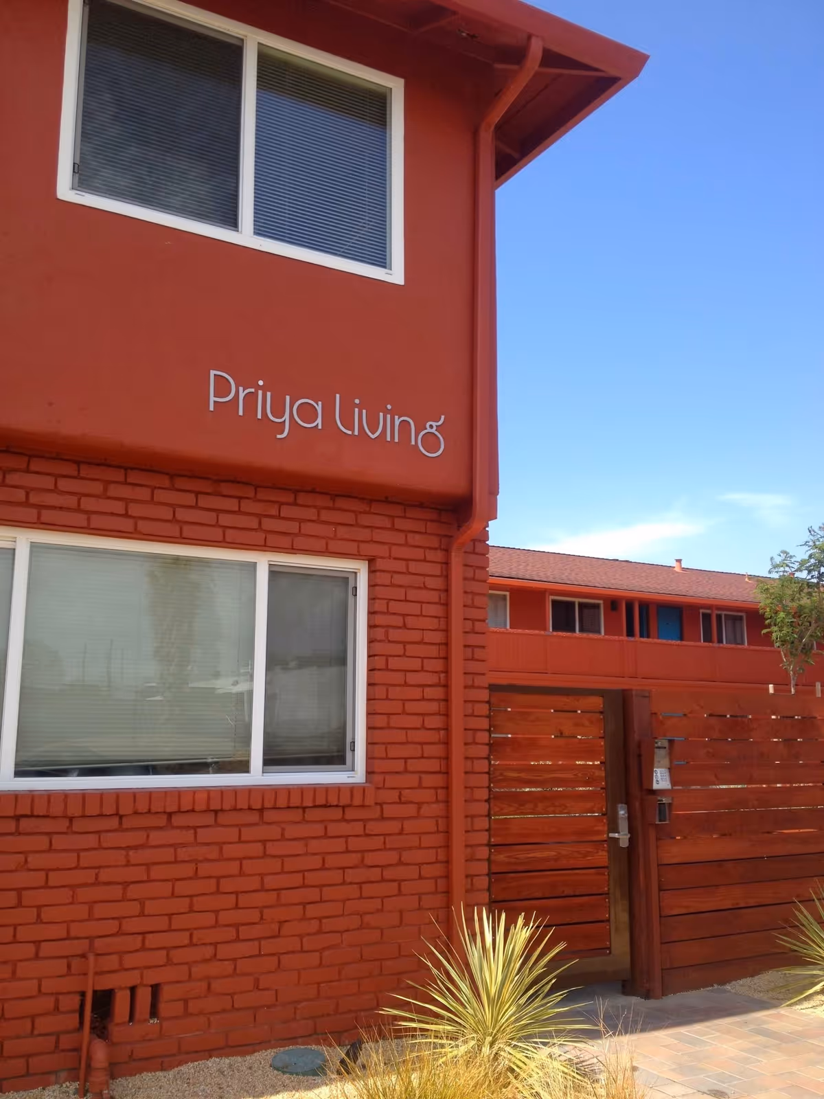 Exterior view of a red brick building with the sign 'Priya Living' on the wall. The building has white-framed windows and a wooden gate with a lock and intercom system. There are some plants in front of the building and a clear blue sky in the background.