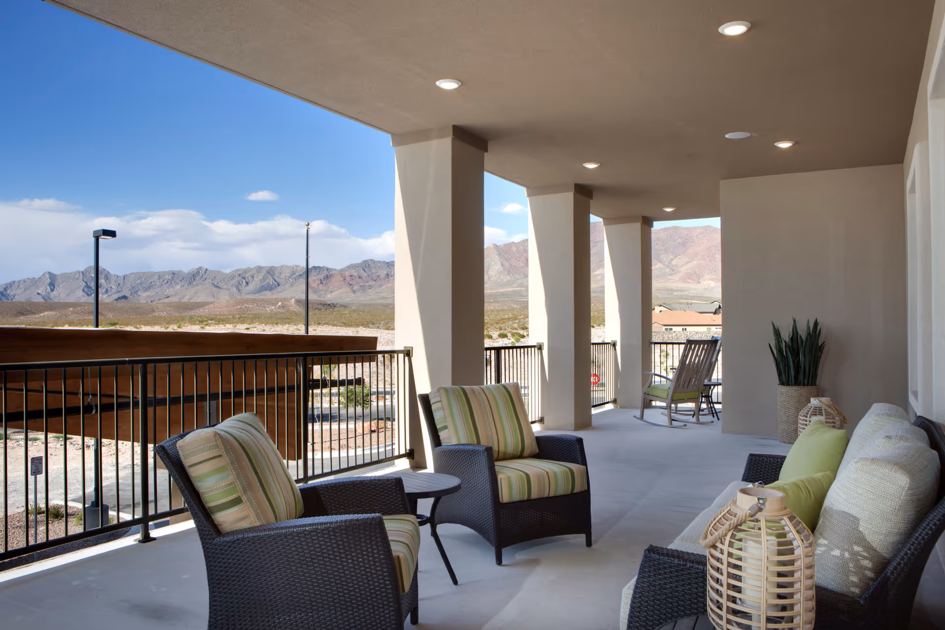 Covered outdoor patio area with cushioned wicker chairs and a sofa, a small table, and decorative lanterns. The patio overlooks a scenic view of mountains and desert landscape under a clear blue sky.