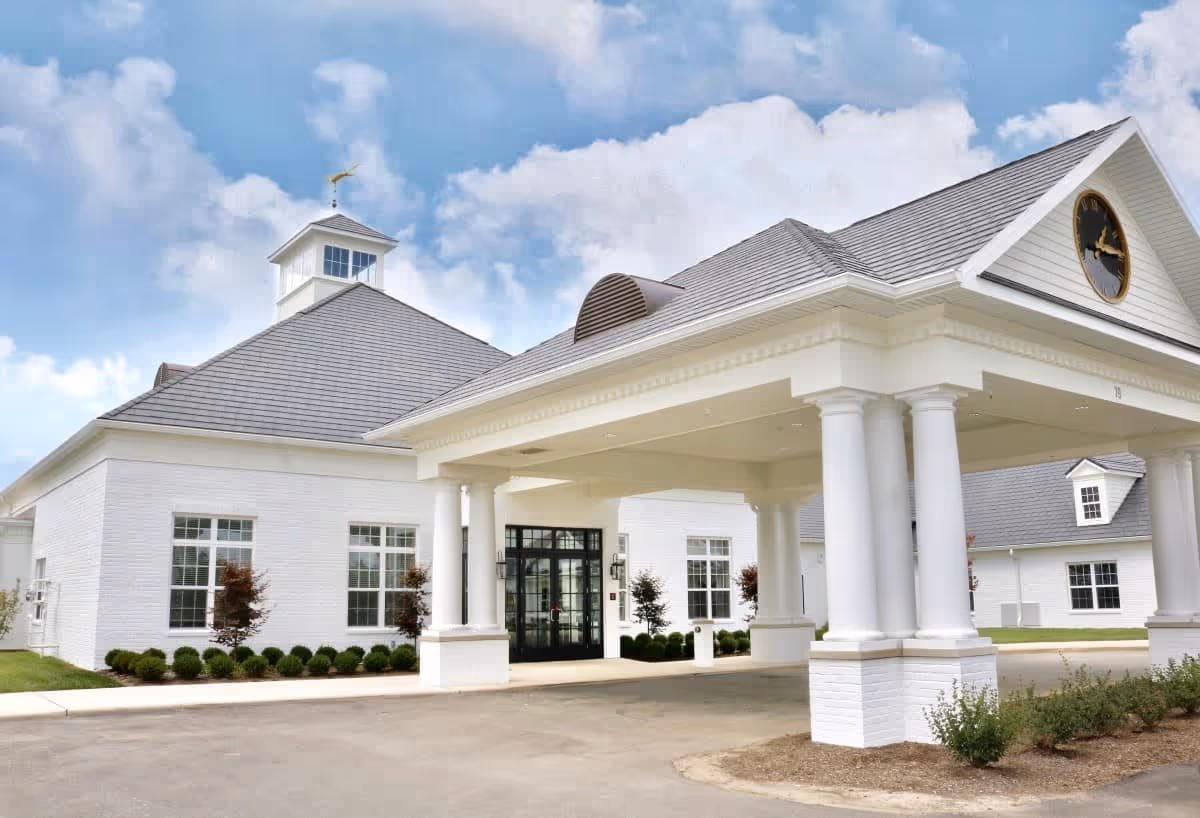 Exterior view of a senior living facility building with white walls, large windows, and a covered entrance supported by white columns. The building has a gray roof with a clock on the front-facing gable and a weather vane on the cupola. The sky is partly cloudy.