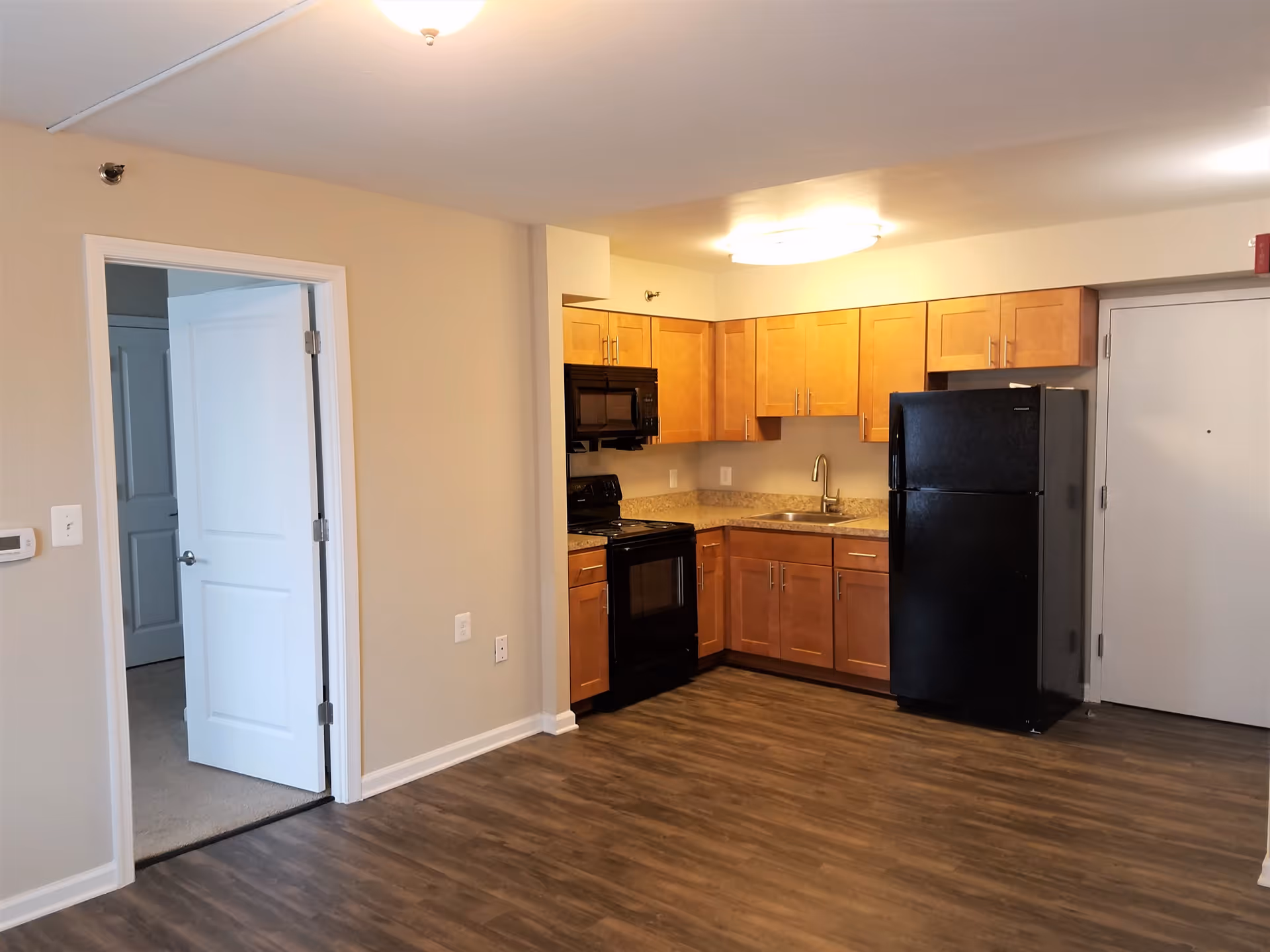 Interior view of a kitchen area in an apartment with wooden cabinets, black appliances including a refrigerator, stove, and microwave. The kitchen has a sink and granite countertops. To the left, there is an open door leading to another room with carpeted flooring. The floor in the kitchen area is wood-style laminate.
