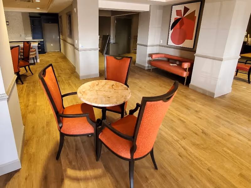 Small communal seating area with a round marble-top table and four orange upholstered chairs on wood flooring, with a bench and artwork on the wall.